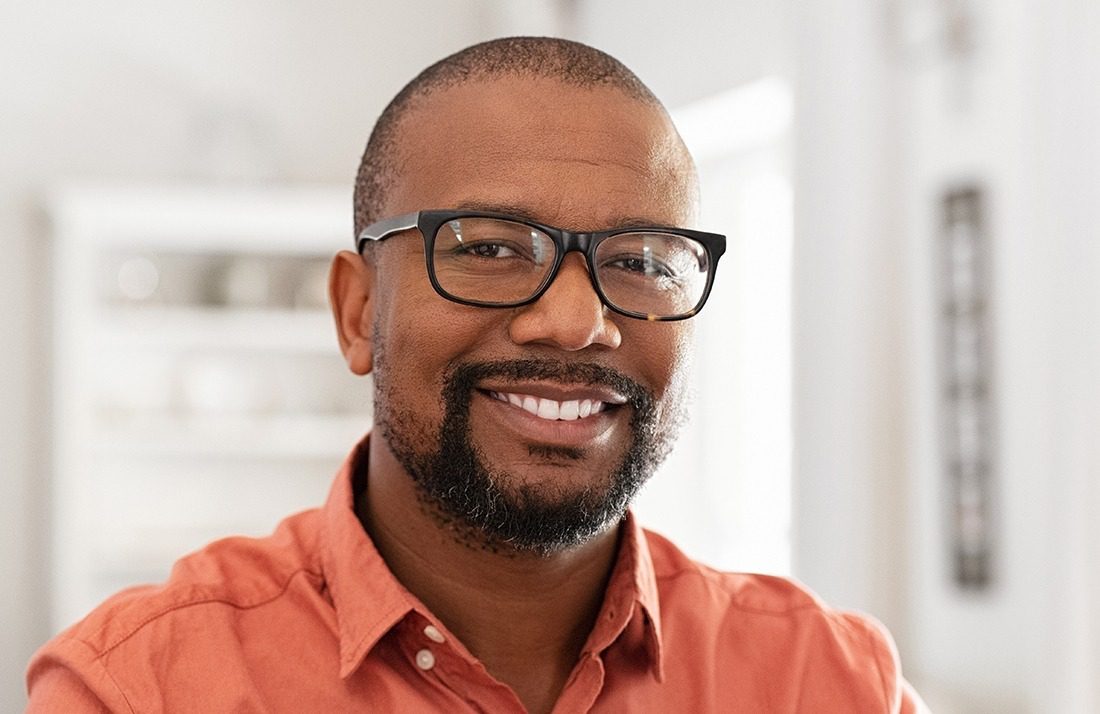 A smiling man with short hair, a beard, and glasses, wearing an orange shirt, stands indoors in a brightly lit room with a blurred background.