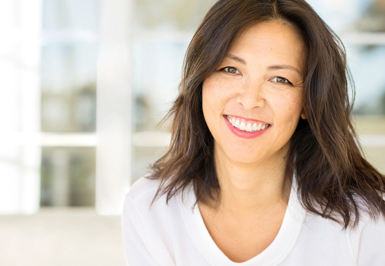 A woman with medium-length dark hair and a white shirt smiling warmly at the camera, with a bright, softly blurred background.