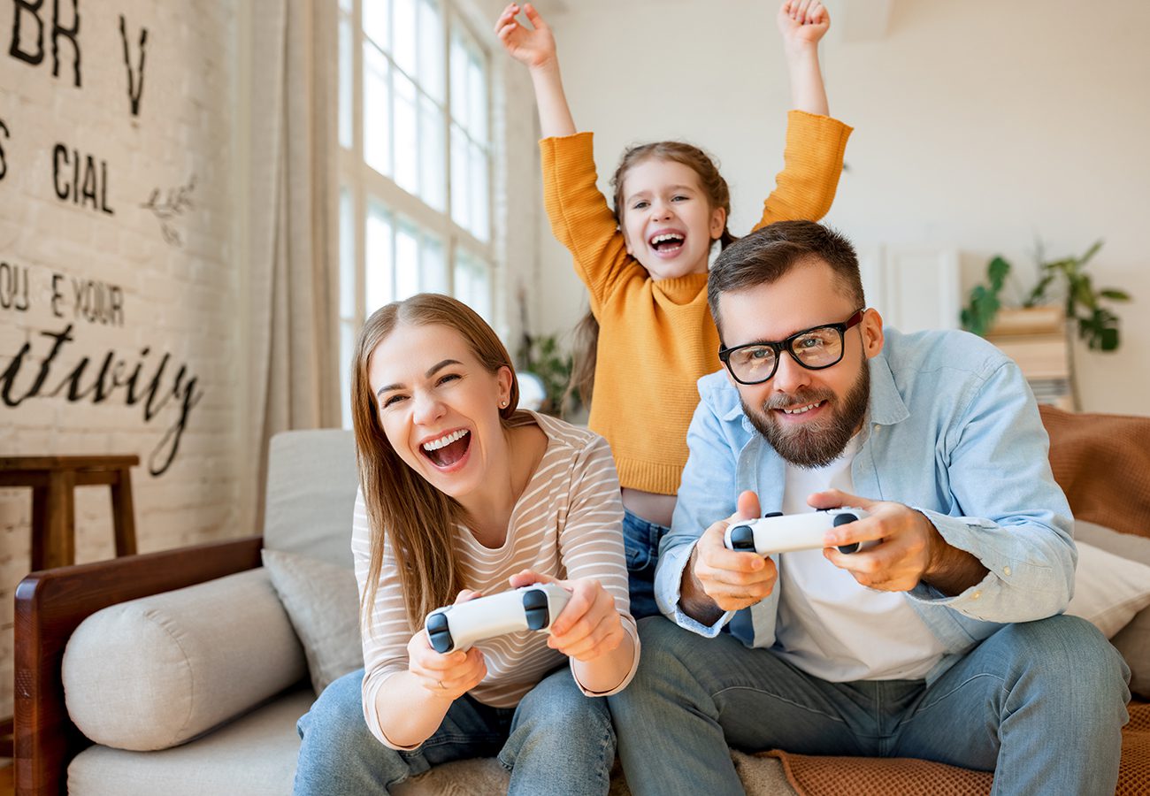 A smiling woman and man sit on a couch playing video games with controllers, while a young girl behind them cheers with her arms raised, all appearing happy and excited in a bright living room.