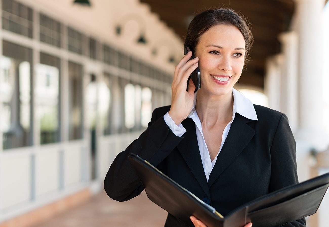 A woman in a business suit smiles while talking on a smartphone. She holds an open black folder and stands in a bright, outdoor corridor with large windows in the background.