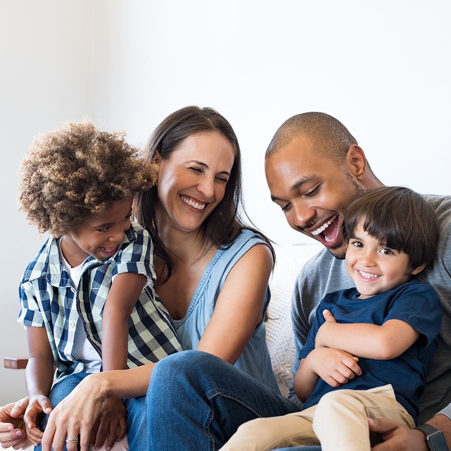 A smiling family of four, including two adults and two young children, sit closely together on a couch, laughing and enjoying each other‚Äôs company in a bright, cozy room.