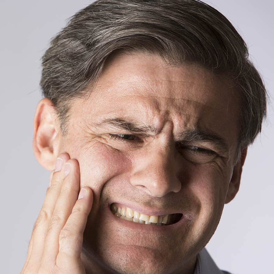 A man with graying hair holds his cheek and winces in pain, suggesting he has a toothache or jaw discomfort.