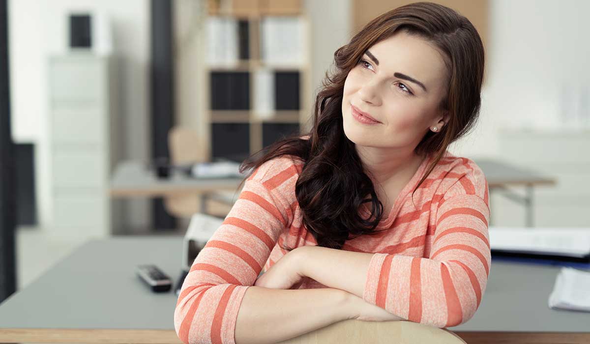 A young woman with long brown hair, wearing a pink and white striped shirt, sits at a desk with her arms crossed on the chair back, smiling and looking thoughtfully to the side in a bright office setting.