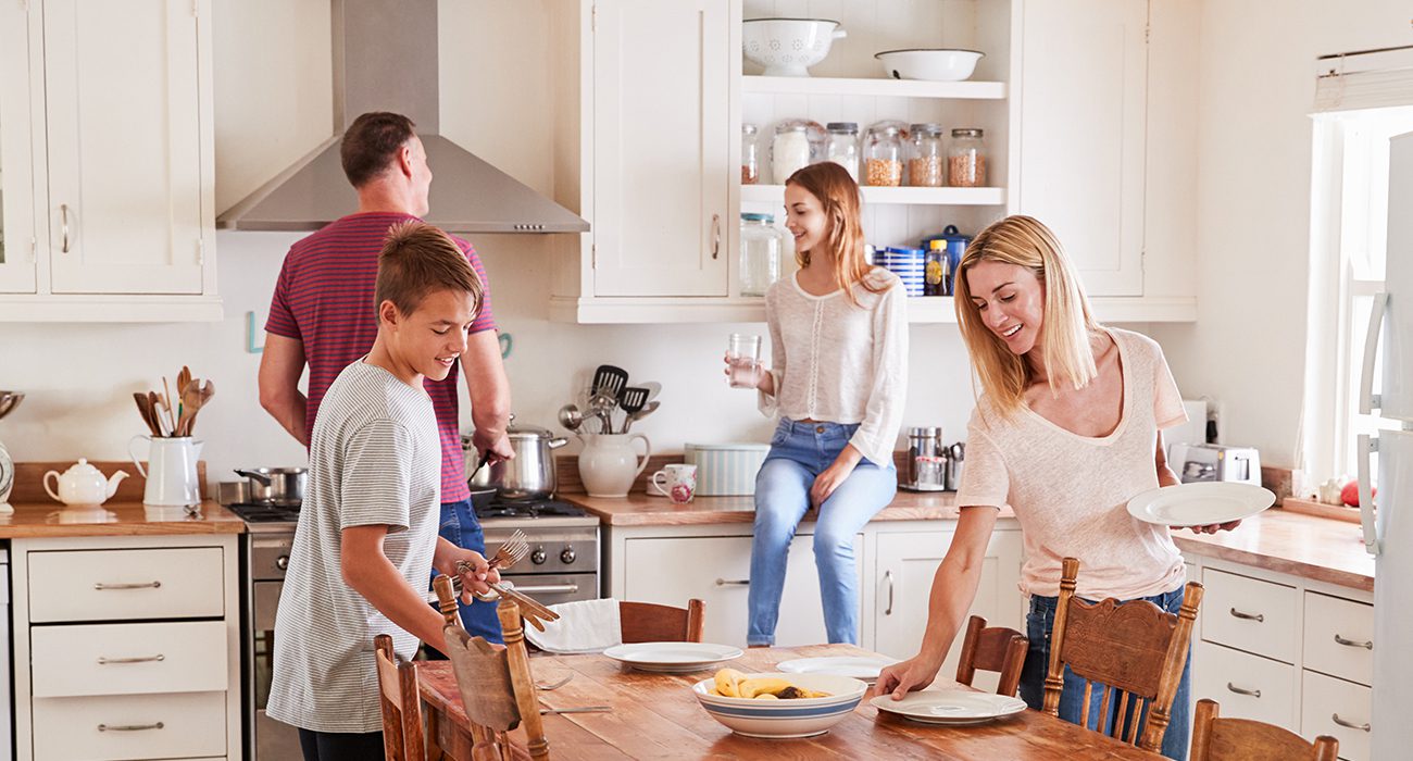 A family of four in a bright kitchen; a woman sets the table, a boy carries cutlery, a man cooks at the stove, and a girl sits on the counter smiling while holding a glass of water.