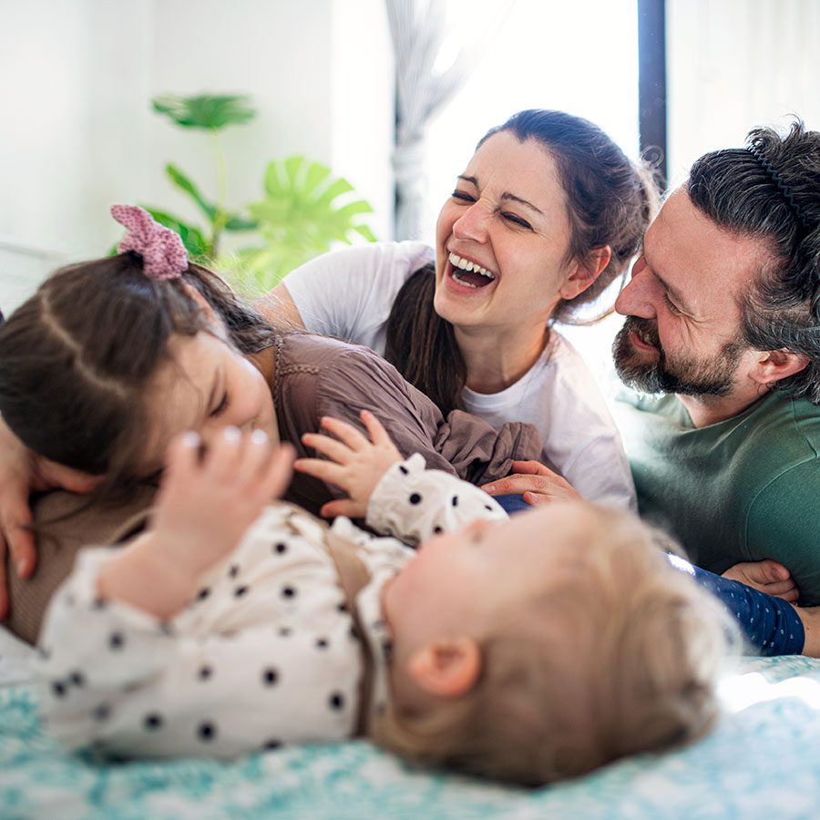 A happy family of four, including two adults and two young children, laugh and play together on a bed in a bright, cozy room. Everyone looks joyful and relaxed.